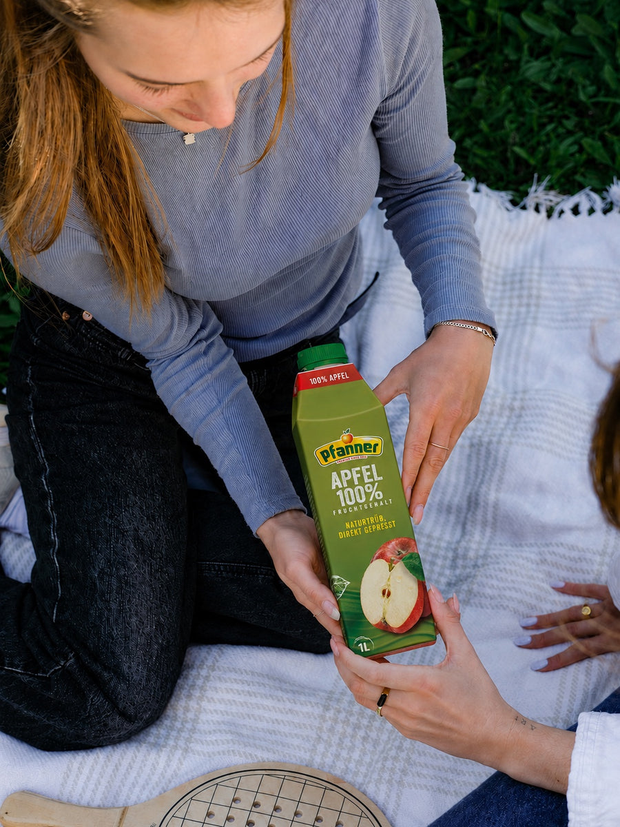 Zwei Personen auf einer Picknickdecke reichen einander eine Packung Pfanner Apfel 100 % Direktsaft